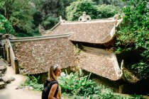 Сверху вид восхитительной женщины-путешественницы с рюкзаком прогуливаясь в тропическом саду рядом с религиозным храмом и наслаждаясь величественным пейзажем — стоковое фото