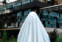 Unerkennbare Person mit weißem Bettlaken maskiert als Gespenst mit rustikalen Metallkonstruktionen auf dem Hintergrund — Stockfoto