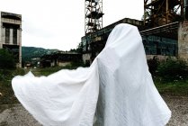 Persona irriconoscibile coperta a mano in lenzuolo bianco mascherato da fantasma con costruzioni in metallo rustico sullo sfondo — Foto stock