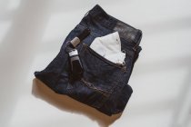 Dall'alto di jeans piegati con maschera protettiva bianca in tasca e chiavi auto posizionate su sfondo bianco in studio leggero — Foto stock