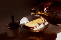 Зверху анонімний врожай виливає оливкову олію з пляшки на хлібний тост, розміщений на столі біля чашки гарячого напою, готуючи сніданок вдома — стокове фото