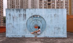 Female in summer dress sitting on concrete base of unusual city installation in shape of passage with round holes and looking away — Stock Photo