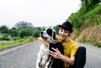 Весела молода леді в повсякденному одязі і капелюсі сидить на землі з мобільним телефоном і приймає селфі з собакою під час прогулянки на вулиці — стокове фото