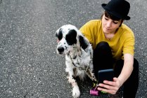 Молода леді в повсякденному одязі і капелюсі сидить на землі з мобільним телефоном і приймає селфі з собакою під час прогулянки на вулиці — стокове фото