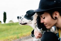 Allegro giovane signora in abiti casual e cappello seduto a terra abbracciando il suo cane durante la passeggiata sulla strada — Foto stock
