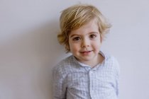 Adorable content child in casual shirt standing near while wall and smiling while looking at camera — Stock Photo