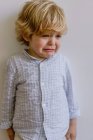 Разочарованный маленький ребенок в повседневной рубашке стоял возле белой стены и плакал на белом фоне в студии — стоковое фото