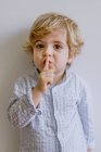 Мила маленька дитина в повсякденному одязі стоїть на білому тлі студії і кладе індексний палець на губи, дивлячись на камеру — стокове фото