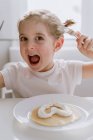 Criança empolgada em camiseta casual sentada à mesa com prato de panqueca saborosa decorada com chantilly em forma de coração e olhando alegremente para a câmera — Fotografia de Stock