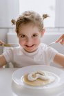Aufgeregtes kleines Kind in lässigem T-Shirt sitzt am Tisch mit einem Teller leckerem Pfannkuchen garniert mit herzförmiger Schlagsahne und schaut fröhlich in die Kamera — Stockfoto