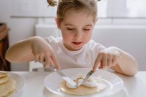 Petit enfant excité en t-shirt décontracté assis à table avec une assiette de crêpe savoureuse garnie de crème fouettée en forme de coeur — Photo de stock