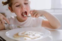 Возбужденный маленький ребенок в повседневной футболке сидит за столом с тарелкой вкусного блина, украшенного взбитыми сливками в форме сердца — стоковое фото