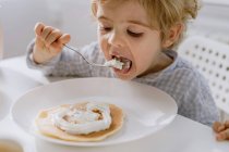 Adorable kid eating delicious pancake garnished with whipped cream while sitting at table in bright kitchen and having breakfast — Stock Photo