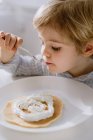 Entzückendes Kind isst leckeren Pfannkuchen garniert mit Schlagsahne, während es am Tisch in der hellen Küche sitzt und frühstückt — Stockfoto