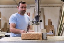 Bajo ángulo de carpintero masculino en ropa casual enfocando y cortando madera usando máquina eléctrica especial mientras trabaja en taller moderno ligero - foto de stock