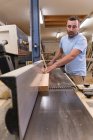 Trabajador de madera masculino en ropa casual enfocando y cortando madera usando una máquina eléctrica especial mientras trabaja en un taller moderno y ligero - foto de stock