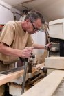 Low angle of attentive middle aged male woodworker in glasses and casual clothes focusing and drilling lumber using special electric machine while working in contemporary workshop — Stock Photo