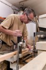Low angle of attentive middle aged male woodworker in glasses and casual clothes focusing and drilling lumber using special electric machine while working in contemporary workshop — Stock Photo