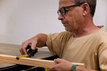 Schreinermeister mit Brille und Freizeitkleidung, der die Größe des hölzernen Details mit Maßband kontrolliert, während er in einer leichten modernen Werkstatt arbeitet — Stockfoto