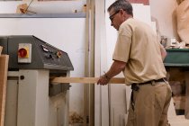Low angle side view of crop attentive middle aged craftsman in glasses and casual clothes focusing and using carpentry electric machine while planing lumber in light modern studio — Stock Photo