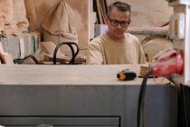 Внимательный работник средних лет в очках и повседневной одежде, фокусирующийся и использующий электрическую машину во время работы с древесиной в современной мастерской. — стоковое фото