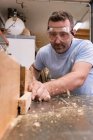 Baixo ângulo de carpinteiro focado em óculos de proteção moagem de madeira enquanto elabora detalhes usando máquina elétrica na oficina moderna leve — Fotografia de Stock