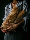 Обрізана невпізнавана жінка в фартусі, що тримає хліб чіабата, стоячи на темному тлі — стокове фото