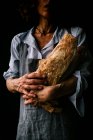 Ritagliato donna irriconoscibile in grembiule in possesso di pane ciabatta mentre in piedi su sfondo scuro — Foto stock