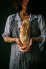 Обрезанная неузнаваемая женщина в фартуке, держащая в руках хлеб чабатта, стоя на тёмном фоне — стоковое фото