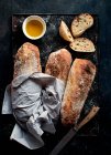 Pan Ciabatta sobre tabla rústica cerca de aceite de oliva y cuchillo con rebanadas de pan sobre fondo oscuro - foto de stock