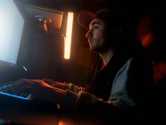 Side view of focused male in cap and wireless headset wearing casual outfit sitting alone at computer and playing video game in dark room with dim blue light at night — Stock Photo