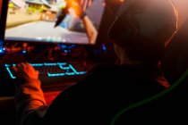 Back view of faceless focused male in cap and wireless headset wearing casual outfit sitting alone at computer and playing video game in dark room with dim blue light at night — Stock Photo