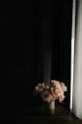 Цветущие свежие розовые розы в винтажной вазе помещены на деревянный стол у черной стены рядом с окнами в современной квартире — стоковое фото