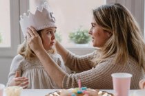 Vista lateral de la alegre madre colocando fieltro corona hecha a mano en la hija mientras celebran cumpleaños juntos en casa - foto de stock