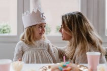 Vista laterale della madre allegra che posiziona la corona fatta a mano in feltro sulla figlia mentre festeggia il compleanno insieme a casa — Foto stock
