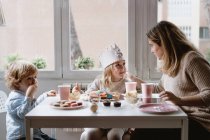 Позитивная мать в повседневной одежде сидит за деревянным столом и разговаривает с детьми, празднующими день рождения дома — стоковое фото