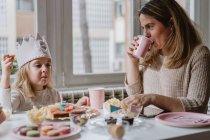 Mãe positiva em roupas casuais sentadas à mesa de madeira e conversando com crianças comemorando aniversário em casa — Fotografia de Stock