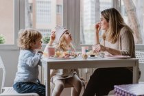 Madre positiva in abiti casual seduta al tavolo di legno e che parla con i bambini che festeggiano il compleanno a casa — Foto stock