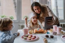 Positive mother and daughter in casual clothes sitting together at table and making video call on tablet while celebrating birthday at home — Stock Photo