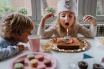 Щаслива маленька дівчинка в повсякденному одязі і з повстяною короною, що дме свічку на день народження торт, сидячи за дерев'яним столом під час вечірки — стокове фото