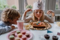 Menina feliz em roupas casuais e com a coroa de feltro soprando vela no bolo de aniversário enquanto sentado na mesa de madeira durante a festa — Fotografia de Stock