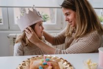 Вид сбоку на веселую мать, надевающую корону ручной работы на дочь во время празднования дня рождения вместе дома — стоковое фото