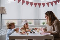 Mère positive en vêtements décontractés assis à la table en bois et parlant avec les enfants célébrant leur anniversaire à la maison — Photo de stock