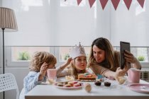 Позитивна мати і дочка в повсякденному одязі сидять разом за столом і роблять відеодзвінки на планшет під час святкування дня народження вдома — стокове фото