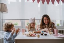 Позитивна мати і дочка в повсякденному одязі сидять разом за столом і роблять відеодзвінки на планшет під час святкування дня народження вдома — стокове фото