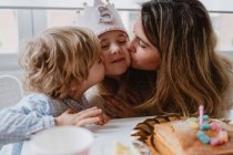 Любляча мати і брат цілуються і вітають маленьку дівчинку разом, проводячи час під час вечірки на день народження за столом вдома — стокове фото