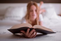 Спокійна жінка розслабляється на ліжку, лежачи на животі і читаючи цікаву книгу в затишній спальні з романтичним написом на стіні і мрійливою атмосферою — стокове фото