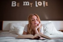 Pacifico feminino relaxante na cama enquanto deitado no estômago e lendo livro interessante no quarto acolhedor com inscrição romântica na parede e atmosfera sonhadora — Fotografia de Stock