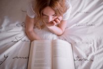 Peaceful female relaxing on bed while lying on stomach and reading interesting book in cozy bedroom with romantic inscription on wall and dreamy atmosphere — Stock Photo