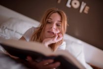 Спокійна жінка розслабляється на ліжку, лежачи на животі і читаючи цікаву книгу в затишній спальні з романтичним написом на стіні і мрійливою атмосферою — стокове фото